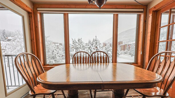 A dining area with a large wooden table and six chairs, large windows with a view of a snowy landscape. - Summit House Short Walk to Skiing, Covered Parking Balcony w/ Panoramic Views (Copper Mountain)