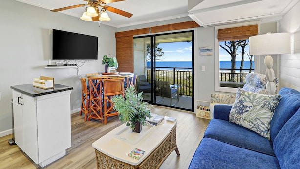 Un salon avec un téléviseur, un coin repas avec des chaises, un balcon avec vue sur la mer et un canapé avec des coussins.
