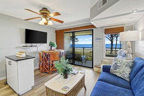 Een woonkamer met een televisie, een eethoek met stoelen, een balkon met uitzicht op zee en een bank met kussens.