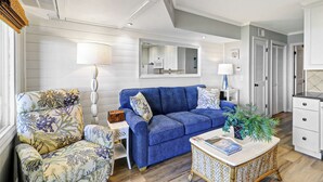 Un salon avec un canapé bleu, des fauteuils à motifs floraux, une table d’appoint avec une lampe, une table basse et un coin cuisine visible à travers une porte ouverte.