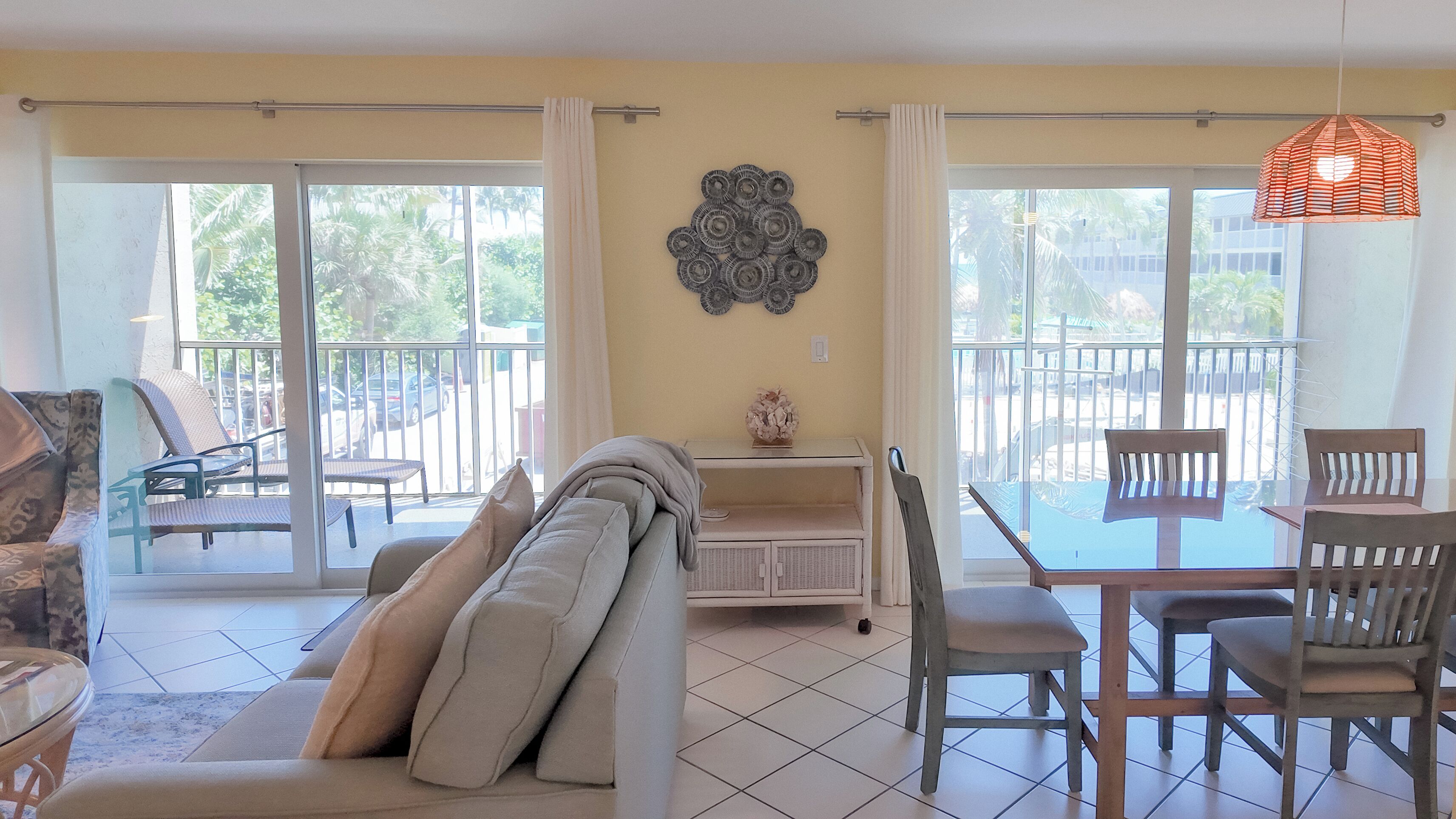A living room with a sofa, chairs, a small table, and a decorative wall piece. There is a sliding glass door leading to an outdoor area with chairs and a table.