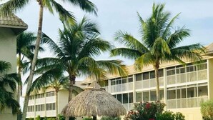 A swimming pool with lounge chairs and a thatched umbrella, surrounded by palm trees and a building with balconies. - Tropical Getaway (Sanibel)
