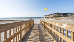Una passerella in legno conduce a una spiaggia con una bandiera gialla, un edificio con un balcone e un cielo sereno.
