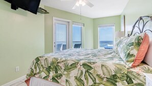 Camera da letto con un letto, una ventola a soffitto, una TV montata a parete e vista sull'oceano dalle finestre.