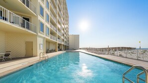 Una piscina adiacente a un edificio a più piani con balconi, cielo sereno e vista sull'oceano in lontananza.