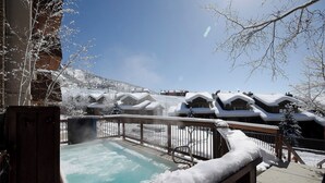 Zona de hidromasaje exterior cubierta de nieve, con una terraza de madera y vistas a edificios nevados al fondo.