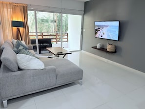 A modern living room with a grey sofa, a small wooden table, a TV on the wall, and a view of a balcony with plants.
