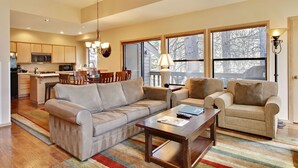 A spacious living room with a sofa, armchairs, and a coffee table. There is a dining area with a table and chairs, and a kitchen with modern appliances. Large windows provide natural light and a view of the outdoors.