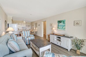 A cozy living room with a sofa, a coffee table, and a dining area with chairs. There is a kitchen in the background with cabinets and a refrigerator.