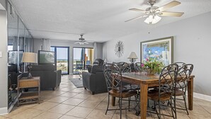 A spacious living area with a dining table, chairs, and a ceiling fan. There is a large window offering a view of the outdoors. - Surf Dweller 102 by Alicia Hollis Rentals FREE TICKETS! $300/Day (Fort Walton Beach)