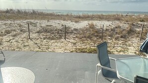 A patio with a glass table and chairs, overlooking a beach with sparse vegetation and a fence. - Surf Dweller 102 by Alicia Hollis Rentals FREE TICKETS! $300/Day (Fort Walton Beach)