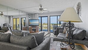 A modern living room with grey sofas, a wooden coffee table, a TV, and a balcony with a view of the beach. - Surf Dweller 102 by Alicia Hollis Rentals FREE TICKETS! $300/Day (Fort Walton Beach)