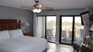 A bedroom with a large bed, a ceiling fan, a nightstand, a lamp, and a view of the outdoors through a sliding glass door. - Surf Dweller 102 by Alicia Hollis Rentals FREE TICKETS! $300/Day (Fort Walton Beach)