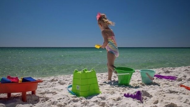 A beach scene with a child playing with sand toys, a green sandcastle, and a red play structure.