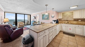 Un'ampia zona giorno con una grande finestra, un divano, una poltrona reclinabile e una cucina con pensili bianchi e un piano di lavoro in marmo.