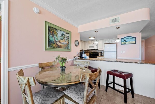 A dining area with a round table, chairs, and a bar stool. A painting hangs on the wall, and a clock is visible. - Island Sunrise 465-Everything is Better in Flip Flops! Book Now! (Gulf Shores)