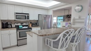 A modern kitchen with white cabinets, stainless steel appliances, a granite countertop, and a breakfast bar with white wicker chairs. - Enjoy Island Living (Sanibel)