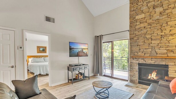 A modern living room with a stone fireplace, a flat-screen TV mounted on the wall, and a large window with a view of greenery. - three bedroom condo with loft, sleeps 8 (Gatlinburg)