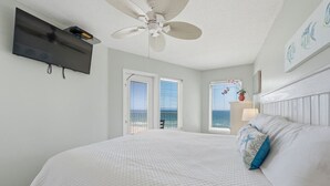 Une chambre à coucher avec un lit, un ventilateur de plafond, une télévision et une vue sur l’océan par la fenêtre.