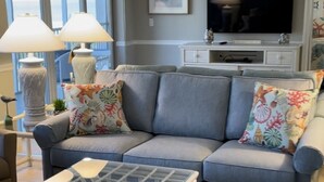 A living room with a grey sofa, floral cushions, a glass coffee table, and a TV on a white cabinet. - Bright And Beachy (Sanibel)