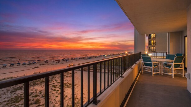 Un balcon avec vue sur une plage au coucher du soleil, des sièges d’extérieur et une balustrade.