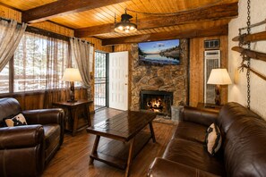 A cozy cabin interior with leather furniture, a stone fireplace, and a television mounted on the wall. - Bear 'n Tree - Walk to Lake & Village, Fireplace, Barbecue (Big Bear Lake)
