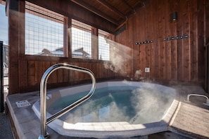 A hot tub with a metal ladder, steam rising, wooden walls, and a view of buildings outside.