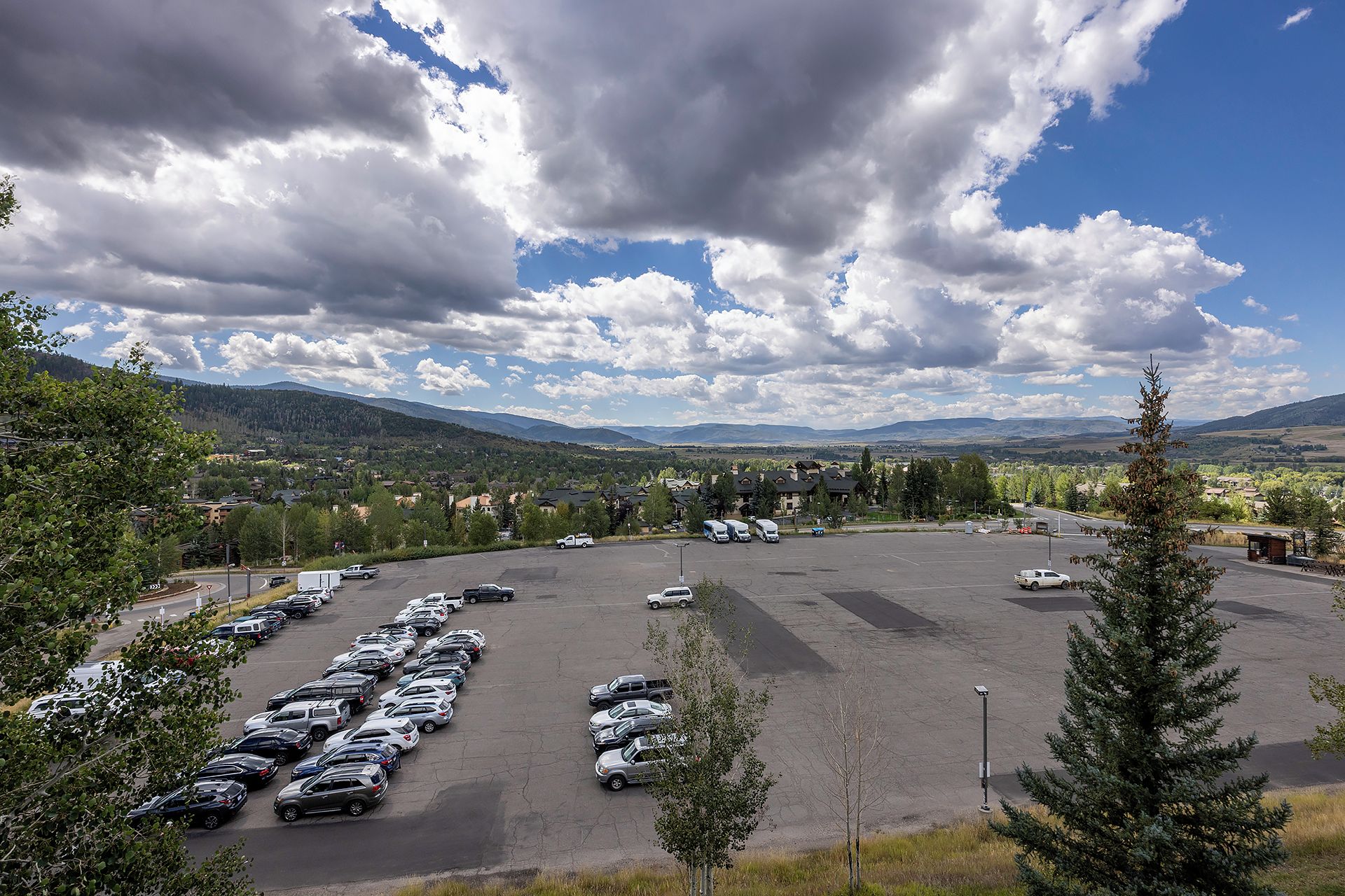 Un parking où sont garées plusieurs voitures, avec vue sur un quartier résidentiel, des maisons et des montagnes en arrière-plan.