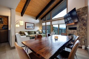 A spacious living area with a wooden dining table, comfortable seating, a flat-screen TV mounted on a stone wall, and large windows offering a view of the outdoors.