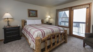 Une chambre à coucher avec un lit en bois, deux tables de chevet avec des lampes, une petite table, une chaise et une grande fenêtre donnant sur un immeuble.