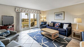 A living room with a TV, sofa, coffee table, and a view of the outdoors through a sliding door. - Stunning Oceanfront Retreat. Steps to the sand and Coligny! (Hilton Head)