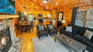 L’intérieur chaleureux d’un chalet avec une cheminée en pierre, un coin repas en bois, une cuisine et un salon doté d’un canapé et d’une table basse.