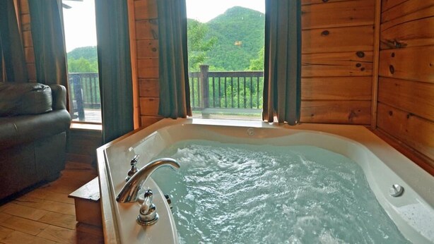 Une cabane en bois avec un jacuzzi, un canapé en cuir et un balcon offrant une vue sur une colline boisée.