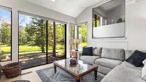 Ein modernes Wohnzimmer mit einem Glastisch, einem grauen Sofa und großen Fenstern mit Blick auf einen grünen Park.