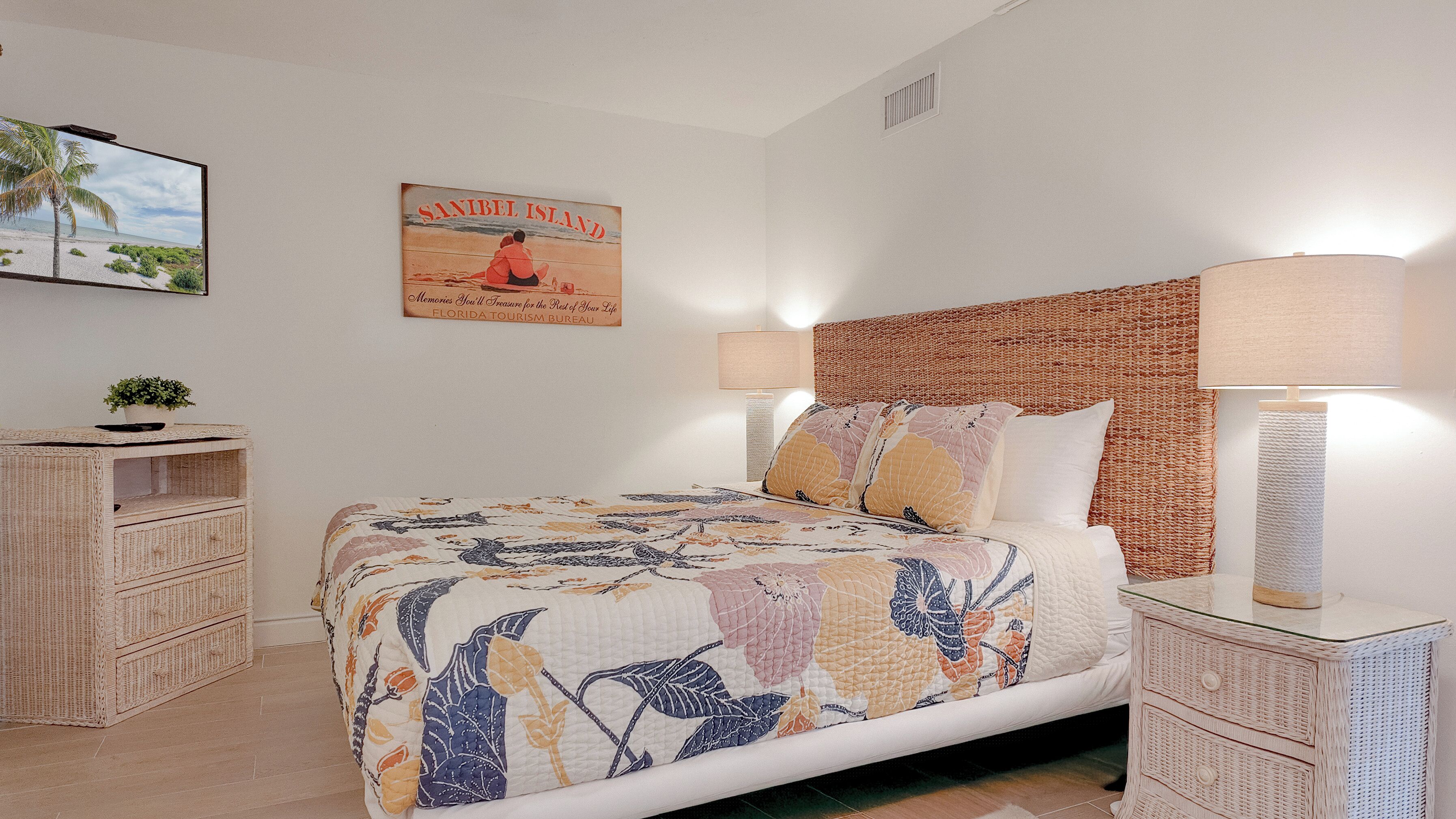 Un dormitorio con una cama, dos mesitas de noche, un televisor montado en la pared, un cartel y una lámpara.