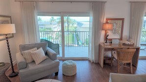A living room with a grey sofa, a round ottoman, a desk with a chair, and a view of a balcony with a pool and palm trees. - WHATA VIEW! (Sanibel)