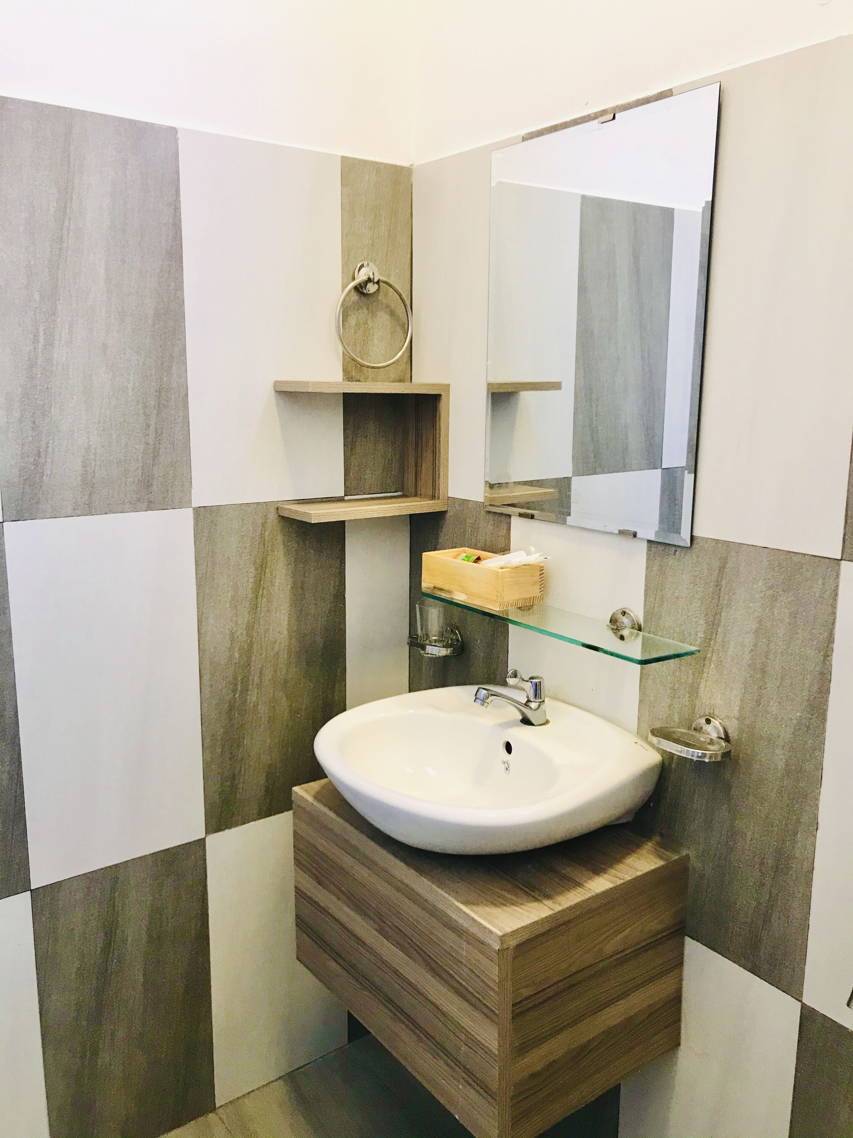 A bathroom with a white sink, wooden vanity, and a mirror above it. There is a towel ring on the wall and a glass shelf with a toothbrush holder.