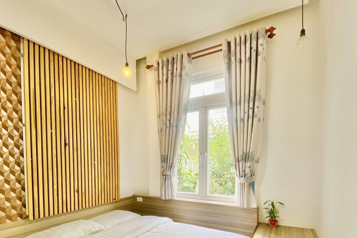 A bedroom with a wooden slatted headboard, a large bed with white bedding, a window with curtains, and a small plant on the windowsill.