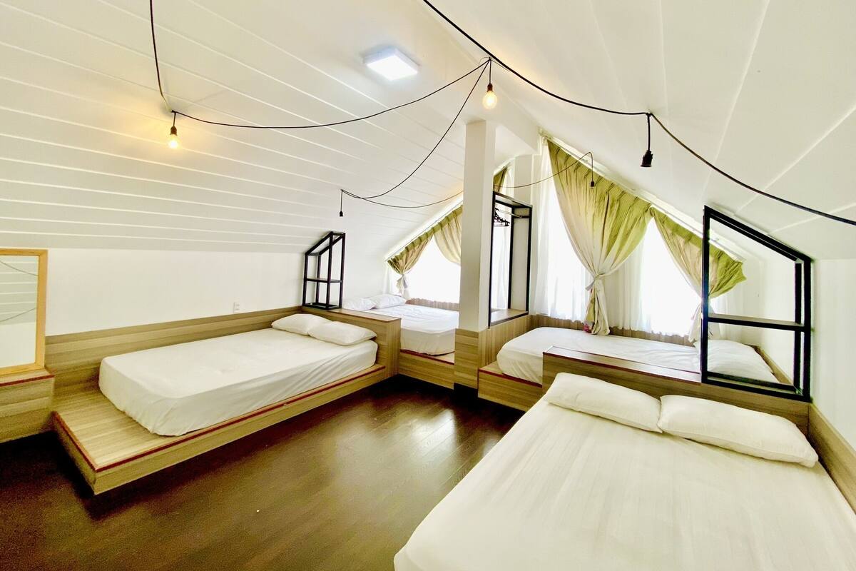 A room with three beds arranged in a U-shape, wooden floors, and a sloped ceiling with exposed beams.
