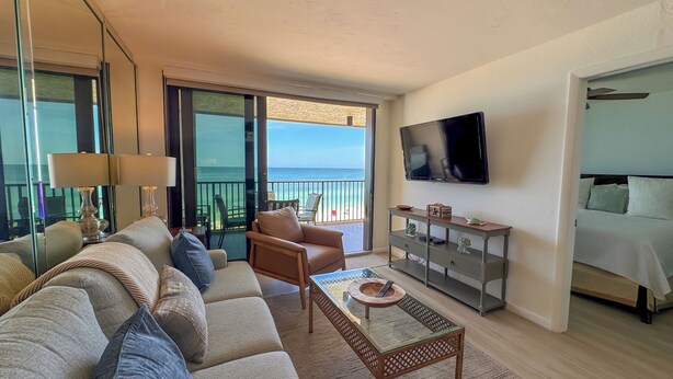 A living room with a sofa, armchairs, a coffee table, a TV, and a balcony with a view of the ocean.