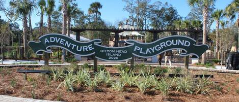 En skylt för Hilton Head Islands äventyrslekplats, med en kant av tegelstenar och omgivande växtlighet.