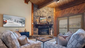 A cozy living room with a stone fireplace, wooden beams, and a large window with blinds. - PX220 | Across the Street from the Slopes | Pool | Hot Tub | Winter Shuttle (Steamboat Springs)