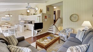 Un salón amplio con un conjunto de sofás, una mesa de centro y un televisor. Hay una zona de cocina con armarios blancos y un microondas. Una escalera conduce a otro nivel.