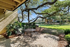 An outdoor patio with a round metal table and chairs, surrounded by lush greenery and a large tree.