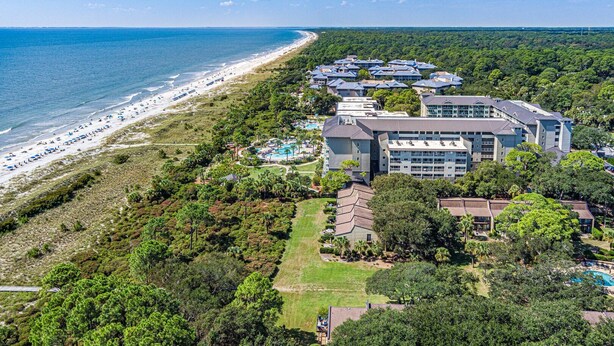 Vista aérea de un complejo turístico frente al mar, con playa, varias construcciones y una piscina.
