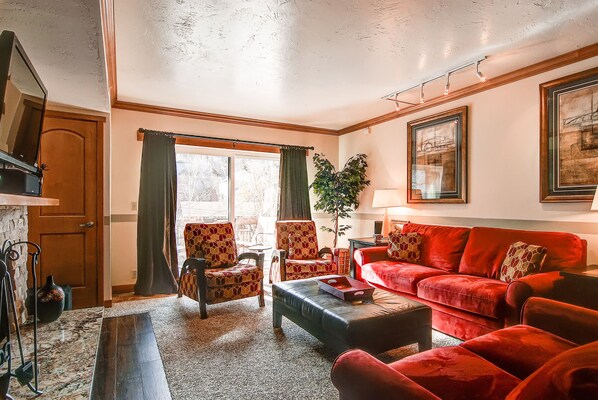 Een woonkamer met een rode bank, gestreept fauteuils, een salontafel en een groot raam met gordijnen.
