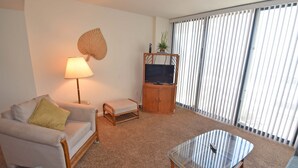 Un salon comprenant un canapé, une table basse en verre, une lampe, un téléviseur posé sur un meuble en bois et une fenêtre avec des stores.