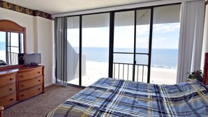 Une chambre à coucher comprenant un lit, une commode, un téléviseur posé sur la commode, et une vue sur la plage grâce à de grandes portes-fenêtres coulissantes.