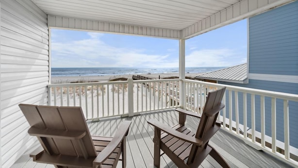 A covered porch with two wooden chairs and a table, overlooking a beach with a clear view of the ocean and sandy shore. - Beach Front Duplex with Gulf Views | Sleeps 11 (Gulf Shores)