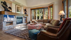 A cozy living room with a stone fireplace, a large window with a view of trees, a leather armchair, and a patterned rug.